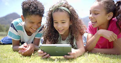 Three biracial children lie on grass, viewing smartphone
