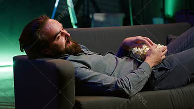 Amused man lounged on couch laughing at funny comedy movie while eating popcorn
