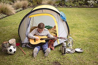 Child boy sitting at backyard tent, strumming guitar with soccer ball and butterfly net nearby