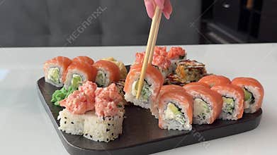 Philadelphia sushi roll picked up with chopsticks from black plate beside California rolls on white table in Japanese