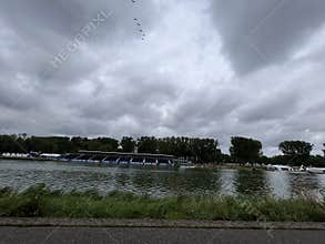 Storm clouds gather above riverside rowing venue with wide water channel, tree