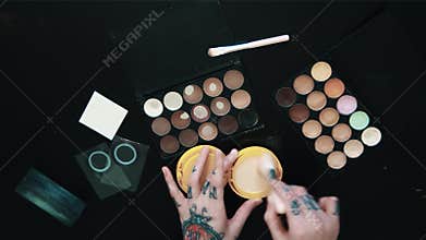 Tattooed female hands uses makeup brush beige powder puff-box over black table