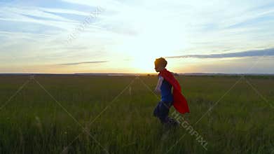 A boy in a superman costume runs across the green field at sunset