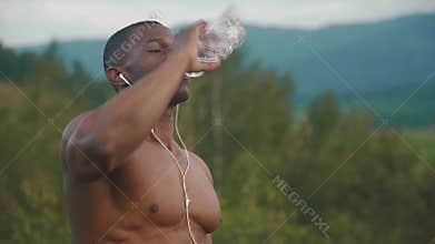 Exhausted african american muscular man with naked torso drinking water after sports training outdoor. Green mountain