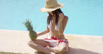 Woman with pineapple and straw hat sits by pool