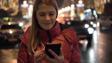 Beautiful attractive young woman standing in a street and using her smartphone. City lights Cars.