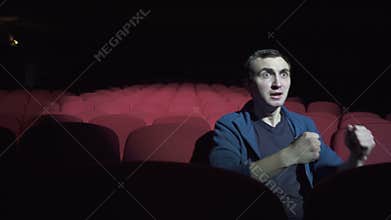 Man sitting in comfortable red chairs in a dark movie theater, watching performance and worried