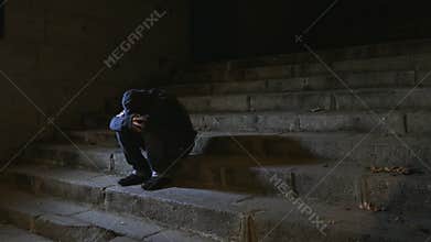 4K video lateral panning 24 fps of young desperate wasted man in hood suffering stress drepression sitting miserable on staircase
