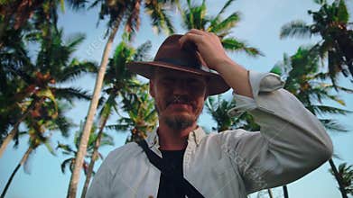 A young active man with a hat with a mustache among the coconut palms wipes sweat from his forehead and then greets the