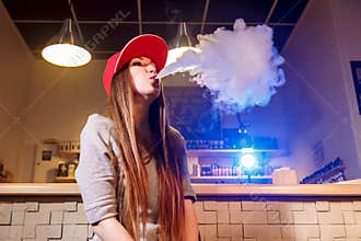 Young pretty woman in red cap smoke an electronic cigarette at the vape shop