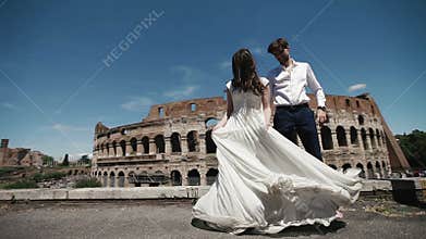 Wedding couple in Rome dancing barefoot at the rooftop near Coliseum, happy groom watches his bride playing with her