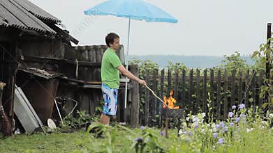 Man with umbrella near the fire for barbecue. The rain is falling, an old building with a fence in the background