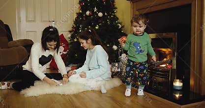 Happy two sisters decorating Christmas and New Year tree and cute son eating apple and looking in camera near fireplace