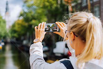 Woman tourist taking a picture of canal in Amsterdam on the mobile phone. Warm gold afternoon sunlight. Travel in Europe