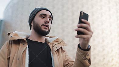 Happy young beard man having video chat holding smartphone webcam chatting with friends. Outside