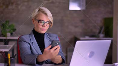 Portrait of middle-aged blonde short-haired businesswoman in glasses making selfie-photo using cellphone in office.