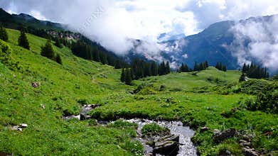 Lush green mountain landscape with meadows and a small creek in the middle under a cloudy and foggy morning sky
