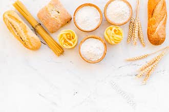 Farinaceous food. Fresh bread and raw pasta near flour in bowl and wheat ears on white stone background top view space