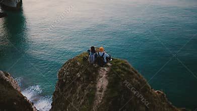 Drone tilts down over happy romantic couple watching sunset ocean view sitting on top of famous Normandy coast cliff.