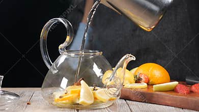 Pouring hot water into teapot with sliced fruit. Glass teapot with orange and lemon inside. Tea brewing Slow motion. Hot