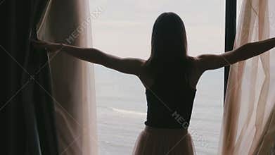 Back view young happy woman walks to open dark hotel apartment room window curtains, enjoy epic sea view slow motion.
