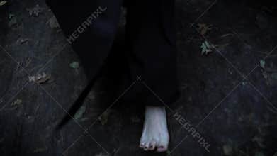 Bare pale feet of woman is wearing long black coat, walking in forest in autumn over wet earth and fallen leaves