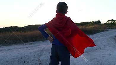 A boy dressed up with a Cape of Superman standing on a mountain looking at the sunset