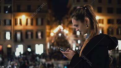 Young attractive woman standing in the city centre at evening and use the smartphone. Crowd and lights on the background