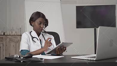 Black female doctor sits at an office desk looking to camera, african american nurse work on laptop in cabinet, modern