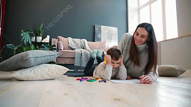 Beautiful young mother in a warm sweater lying on the floor with my son drawing with markers on paper portraying his
