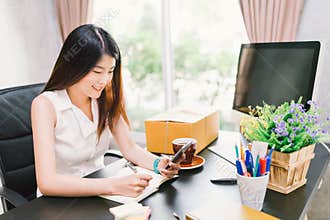 Asian small business owner work at home office, using mobile phone call, writing confirm purchase order on notebook