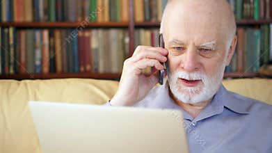 Senior man sitting at home with laptop and smartphone. Using cellphone discussing project on screen