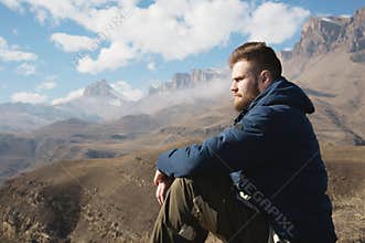 A portrait of a stylish bearded hipster sitting on a rock against the backdrop of epic rocks and contemplating into the