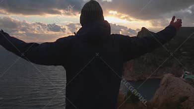 Silhouette of Man in Jacket and Cap Standing with Raised Hands and Looking at Sunset