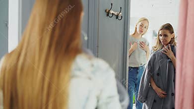 Friendly saleswoman is helping female customer to choose a coat. Cheerful shopper is standing in fitting room in front