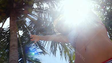 Bottom of view of young woman in bikini dances stands under the palm tree with coconuts on the sun on the