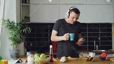 Cheerful happy man dancing and singing in kitchen while surfing social media on his smartphone at home in the morning