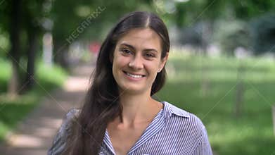 Portrait of charming young woman looking and smiling with teeth at camera, standing in park, daytime, outdoors