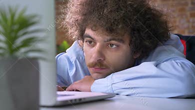 Bored nerdy boss with curly hair and mustache lying on table and working on laptop, modern office background