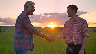 Old farmer greeting young businessman and shaking hands, standing on wheat field, beautiful nature with sunset in