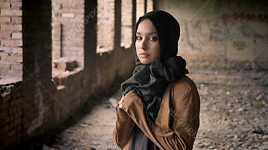 Young beautiful muslim woman in black hijab standing in abandoned building and looking at camera with scared and
