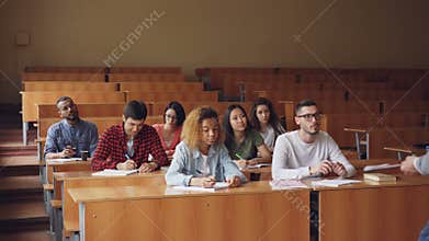 Diligent students are writing making notes in notebooks sitting at lecture at college while teacher is speaking standing