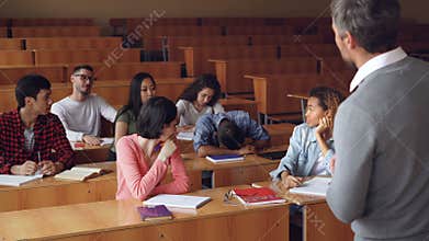 Tired mixed race student is sleeping during lecture at college, his classmates are waking him up, he is standing and
