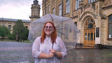 Fat ginger girl with glasses is standing in park in rainy weather, holding umbrella, watching at camera, smiling
