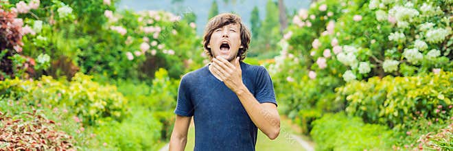 Young man sneeze in the park against the background of a flowering tree. Allergy to pollen concept BANNER long format