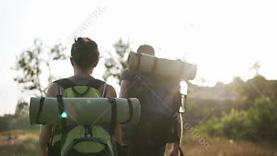 Two travelers - man and woman with huge backpacks are hiking. Walking by grass hills. Sun shines on the background