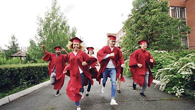 Dolly shot of excited grads running on campus wearing gowns and traditional hats celebrating end of studies. Higher
