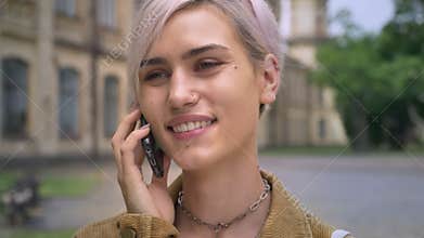 Young happy blonde woman with piercing and short hair talking on phone and smiling, standing near university