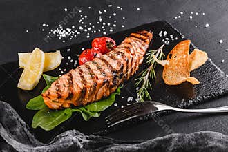 Grilled salmon fillet garnished with spinach, lemon, herbs on plate over wooden background. Hot fish dish