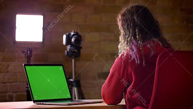 Rear view of young blogger in headphones recording video on camera in front of laptop on bricken wall background.